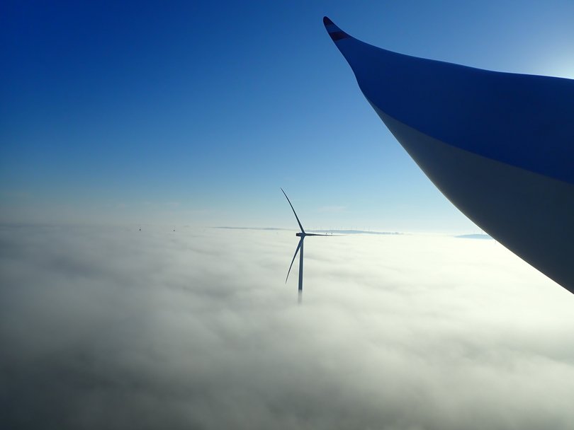 Windräder ragen aus einer dichten Nebeldecke in den blauen Himmel, im Vordergrund eine Rotorblattspitze.